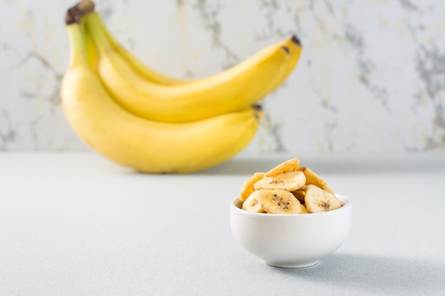 Batatas fritas de banana assada em uma tigela branca e um cacho de bananas na mesa.