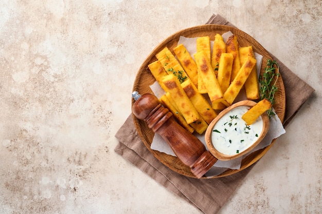 Batatas fritas de polenta caseiras com sal marinho, parmesão, tomilho, alecrim com molho de iogurte. Polenta frita italiana típica. Palitos de milho frito. Antigo fundo claro de concreto. Vista do topo.