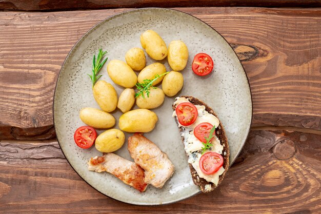 Batatas jovens cozidas, pedaços de carne de peru grelhada e pão de cereais com queijo Roquefort e tomate em um prato sobre um fundo de madeira.