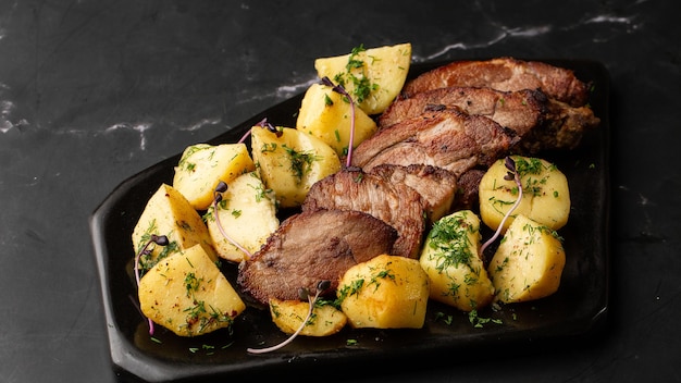 Batatas novas assadas no forno com sal marinho e lombo de porco cortado em pedaços em uma placa preta na mesa de mármore