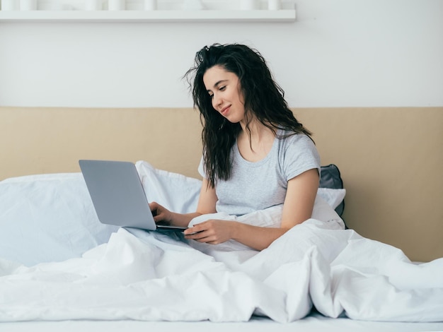 Bate-papo matinal Comunicação online Tecnologia moderna Lazer em casa Mulher feliz relaxada gostando de ler mensagem usando laptop na cama no quarto claro