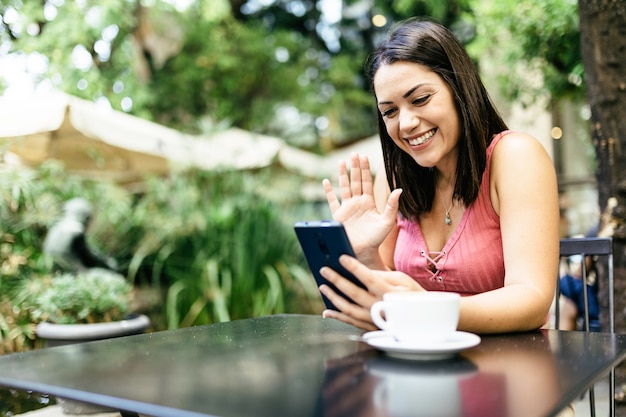 Foto bate-papo por vídeo online - mulher jovem hispânica sorridente usando telefone inteligente em um café e conversando com amigos por meio de aplicativos para dispositivos móveis em redes sociais - conceito de estilo de vida influenciador e vlogger