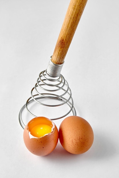 Foto batedor espiral para bater e dois ovos de galinha bege em um fundo branco cozinhando uma omelete