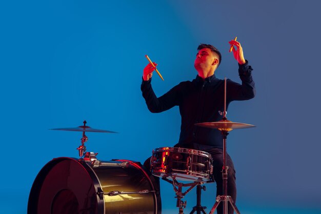 Baterista caucasiano improvisando isolado no fundo azul do estúdio sob luz de néon