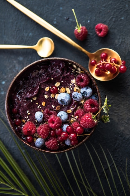 Batido de acai congelado en cáscara de coco con frambuesas, plátano, arándanos, frutas y granola sobre fondo de hormigón. Desayuno, comida saludable para las vibraciones del verano, vista superior, espacio para texto