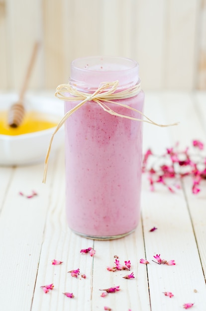 Foto batido cor-de-rosa da morango no frasco retro com flores roxas e mel em placas rústicas claras.