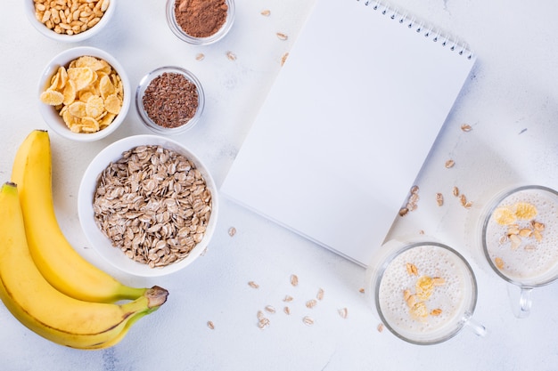 Batido de banana em xícaras, sementes de linho, cacau, aveia e arroz tufado em um fundo branco. Conceito de alimentação saudável