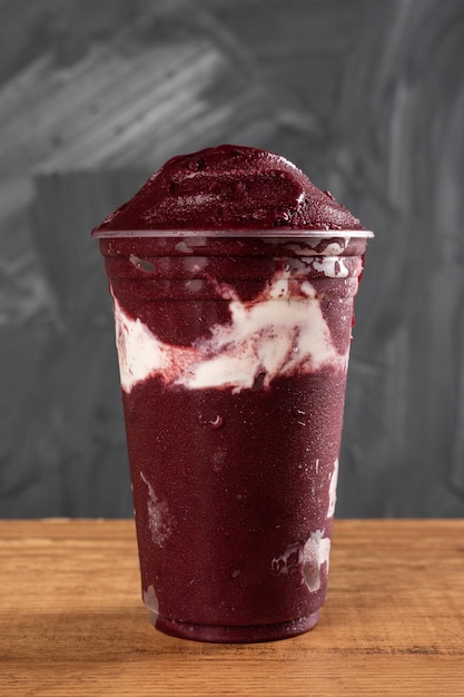 Batido de Sorvete Brasileiro Frozen AÃ§ai Berry em copo plÃ¡stico com Leite Condensado. Sobre uma mesa de madeira e um fundo cinza de verão. Vista frontal para menu e mídia social