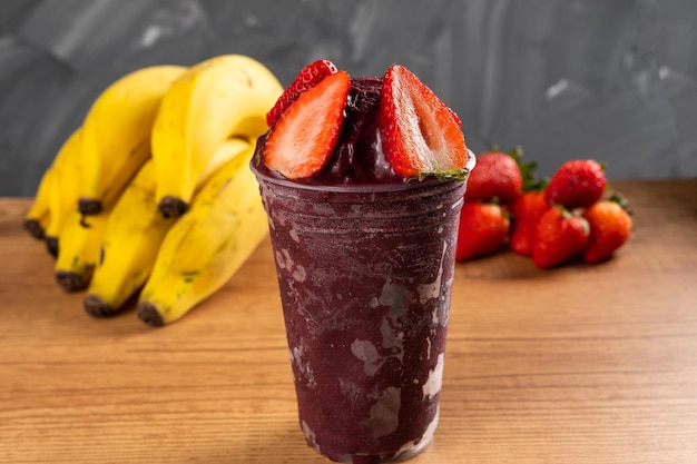 Batido de sorvete brasileiro frozen aã§ai berry em copo plã¡stico com morangos. sobre uma mesa de madeira e um fundo cinza de verão com frutas. vista frontal para menu e redes sociais.