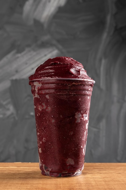 Batido de helado brasileño congelado AÃ§ai Berry en vaso de plástico. Sobre un escritorio de madera y un fondo gris de verano. Vista frontal para menú y redes sociales.