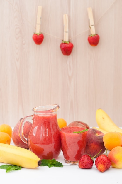 Batido de plátano fresa saludable con menta en un vaso y jarra sobre fondo de madera. Fondo de plátanos, duraznos y albaricoques de frutas frescas