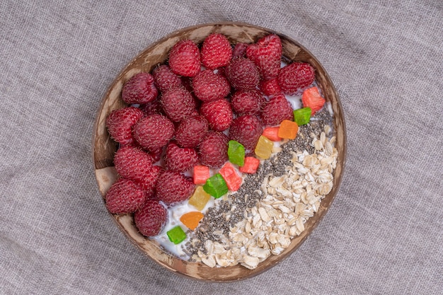 Batido en tazón de coco con frambuesas, avena, frutas confitadas y semillas de chía para el desayuno, de cerca. El concepto de alimentación saludable, superalimento. Vista superior