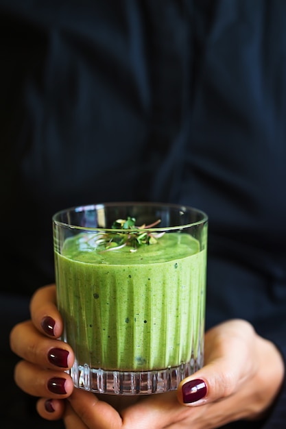 Batido vegano verde con espinacas, plátano y semillas germinadas en vidrio en manos femeninas sobre fondo negro Concepto de desintoxicación de alimentación limpia