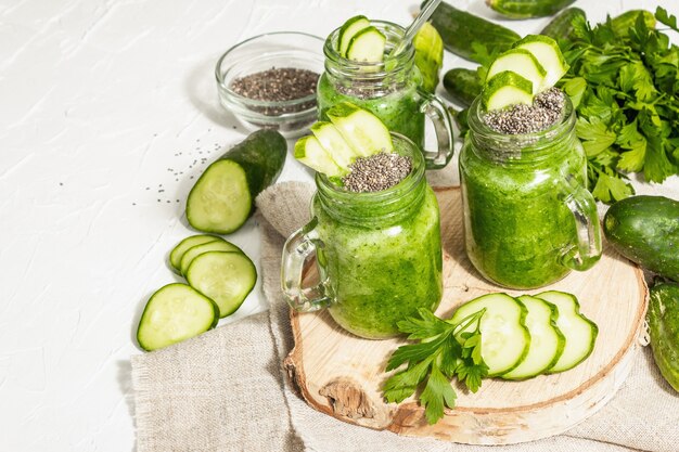 Batido verde con pepino en un frasco de vidrio. Verduras frescas maduras, verduras y semillas de chía. Luz dura de moda, sombra oscura. Fondo de masilla blanca, cerrar