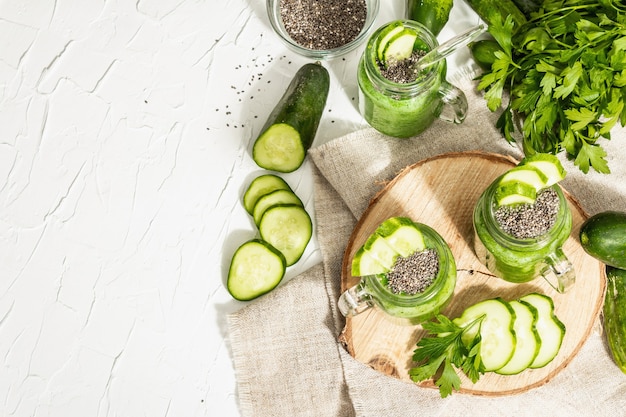 Batido verde con pepino en un frasco de vidrio. Verduras frescas maduras, verduras y semillas de chía. Luz dura de moda, sombra oscura. Fondo de masilla blanca, vista superior