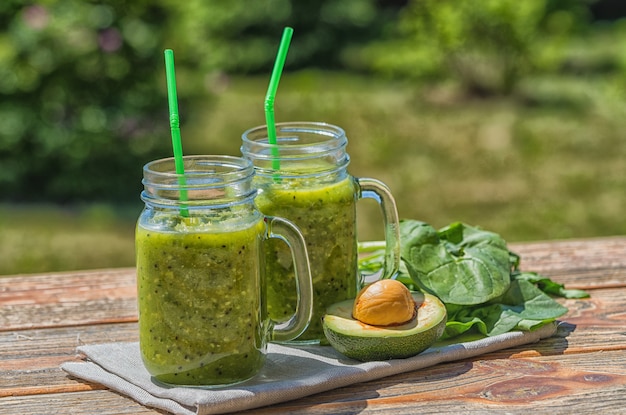 Batido verde vegano saludable con espinacas en frascos de vidrio y aguacate, al aire libre, sobre fondo de naturaleza.