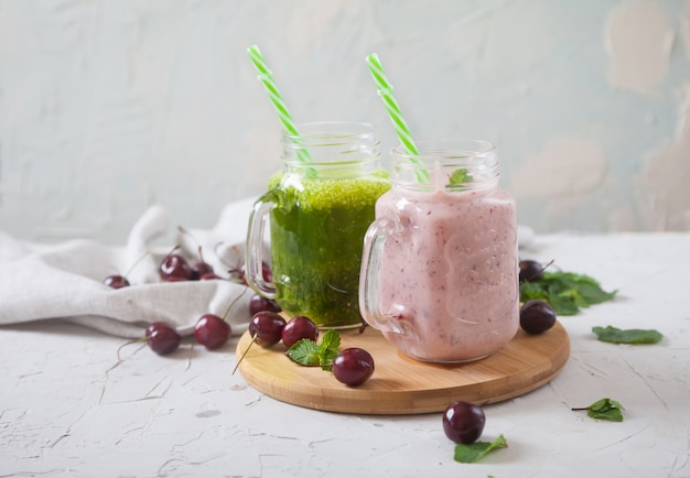 Batidos de cereza verde y dulce sobre un soporte de madera, cerezas esparcidas y una servilleta de lino sobre un fondo de hormigón.