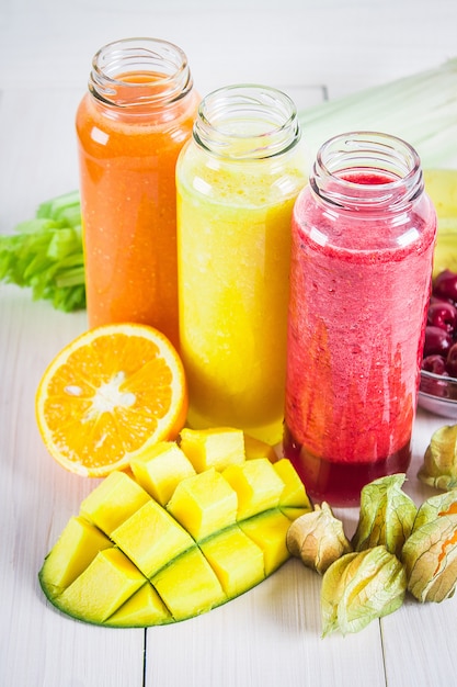 Batidos multicolores en botellas de mango, naranja, plátano, apio, bayas, en una mesa de madera.