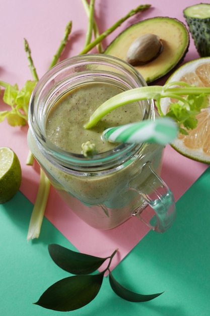 Batidos vegetarianos saludables de verduras con hojas verdes, rodajas de limón, aguacate, cocumber y paja de plástico en un recipiente de vidrio sobre fondo de papel verde rosa duotono.