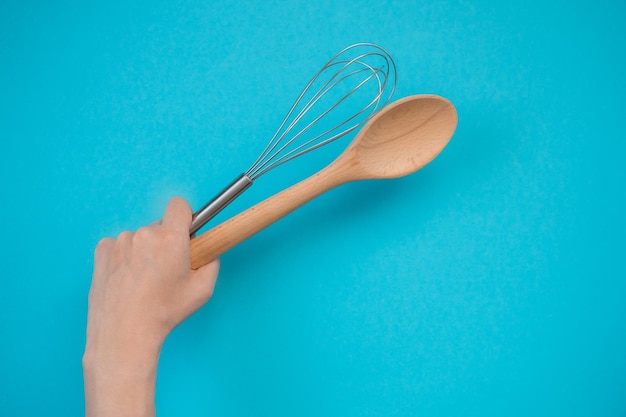 Batir y una cuchara de madera en una mano femenina sobre un fondo azul Lugar para texto o información