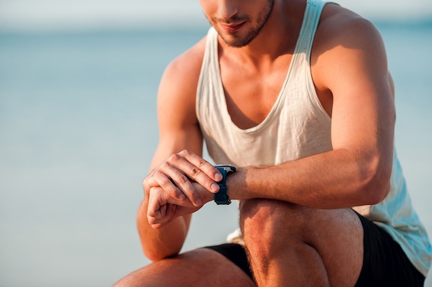 Va a batir su mejor tiempo. Imagen recortada de un joven musculoso que controla el tiempo en sus relojes mientras está sentado al aire libre