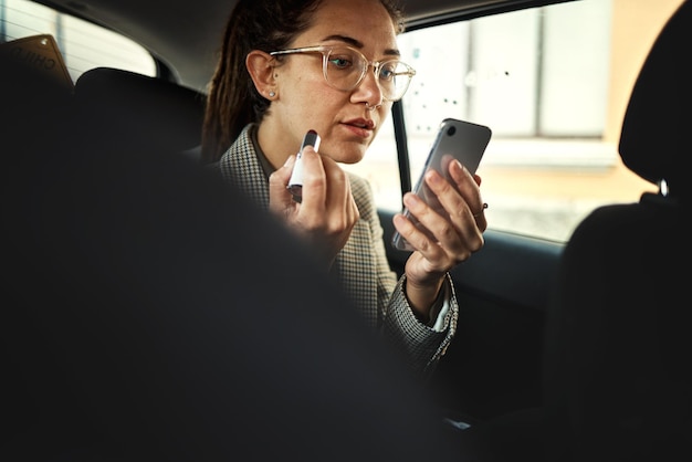 Batom de telefone e uma mulher de negócios em um táxi para viajar na cidade para trabalhar enquanto multitarefa Face móvel e maquiagem com uma jovem funcionária em um carro ou táxi como passageira para transporte