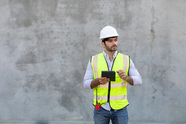 Bauarbeiter, der digitales Tablettenisolat auf grauem Zementhintergrund hält und betrachtet. Projektingenieur auf der Baustelle.