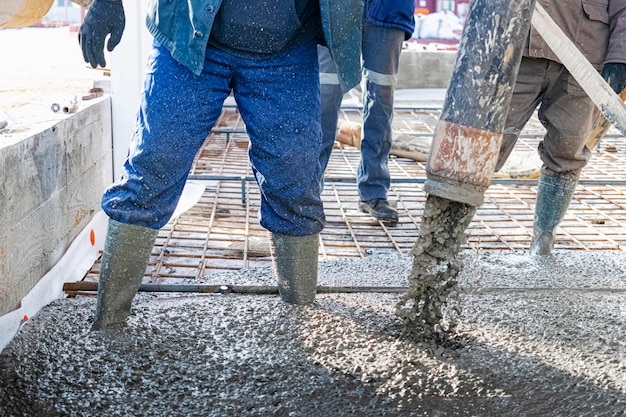 Bauarbeiter gießen Betonboden in Industriewerkstatt. Beine in Stiefeln in Beton. Vorlage von Beton zum Gießen des Bodens. Monolithische Betonarbeiten.