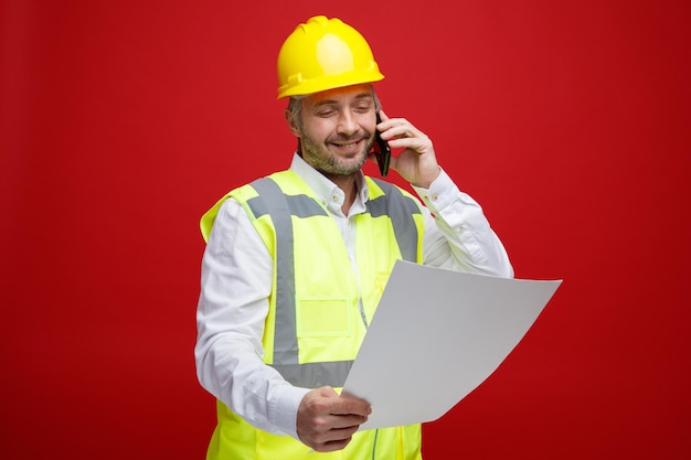Bauarbeiter in Bauuniform und Schutzhelm mit einem Plan, der ihn mit einem Lächeln im Gesicht betrachtet und mit dem Handy spricht, das über rotem Hintergrund steht