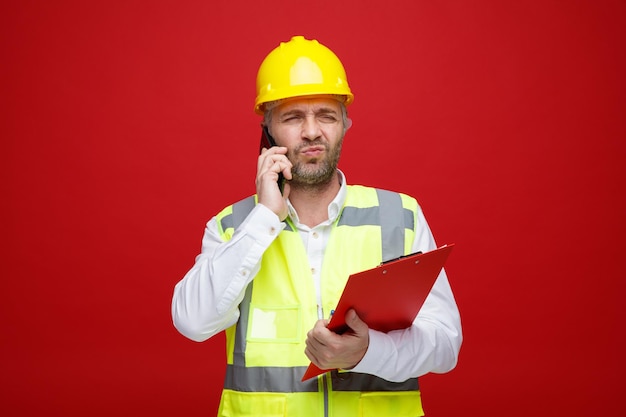 Bauarbeiter in Bauuniform und Schutzhelm mit Klemmbrett am Handy sprechend unzufrieden stirnrunzelnd vor rotem Hintergrund stehend