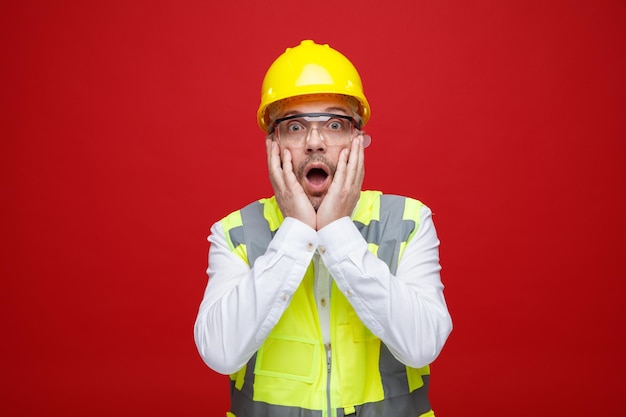 Foto bauarbeiter in bauuniform und schutzhelm mit schutzbrille, der erstaunt und überrascht in die kamera blickt und händchen auf hicksbacken hält, die über rotem hintergrund stehen