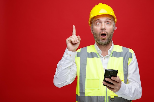 Bauarbeiter in Bauuniform und Schutzhelm mit Smartphone, der erstaunt und überrascht in die Kamera blickt und den Zeigefinger über rotem Hintergrund zeigt