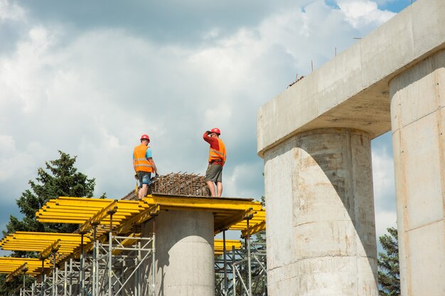 Bauarbeiter mit Schutzhelmen montieren beim Bau einer Straßenbrücke verstärkte Rahmen auf Betonsäulen.