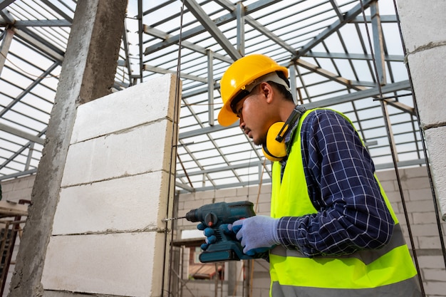 Bauarbeiter verwenden einen Bohrer, Ingenieur, der Sicherheitsausrüstung (Helm und Jacke) trägt, verwendet eine Bohrmaschine, um eine belüftete Mauer zu montieren.