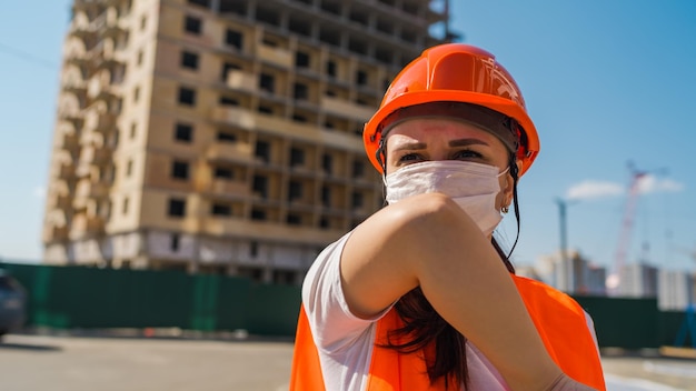 Bauarbeiterin in Overalls und medizinischer Maske, die im Ellbogen auf dem Hintergrund des im Bau befindlichen Hauses hustet Konzept der Bedrohung durch eine Coronavirus-Epidemie-Infektion