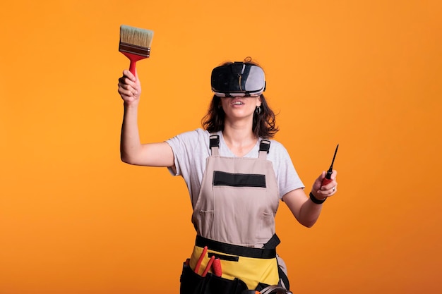 Bauarbeiterin mit Vr-Brille, während sie Pinsel im Studio hält. Bauunternehmerin mit Overalls mit Virtual-Reality-Headset vor dem Streichen von Wänden für Renovierungsprojekte.