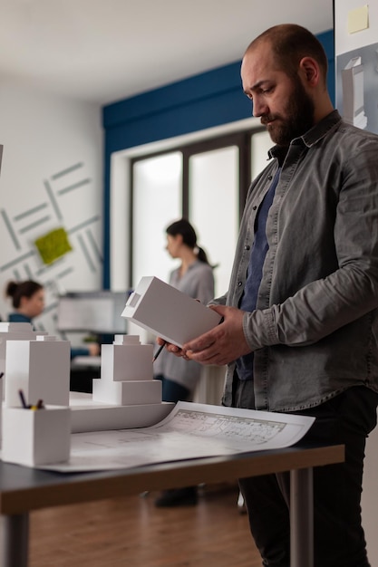 Bauarchitekt, der Schaummachette in den Händen hält und nach Inspiration sucht, steht neben dem Schreibtisch mit Blaupausen. Architekturingenieur im modernen Büro vergleicht Baupläne mit 3D-Modell.