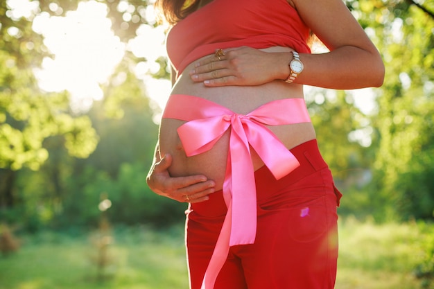 Bauch der schwangeren Frau mit einem rosa Band gebunden. Porträt der schwangeren Frau im Freien im sonnigen Tag mit Platz für Text bewältigen. nackter Bauch der Frau, die ein Baby erwartet. 9 Monate gesunde Schwangerschaft