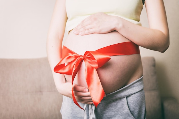 Bauch einer schwangeren Frau, die mit einer roten Schleife auf einem häuslichen Hintergrund gefesselt ist