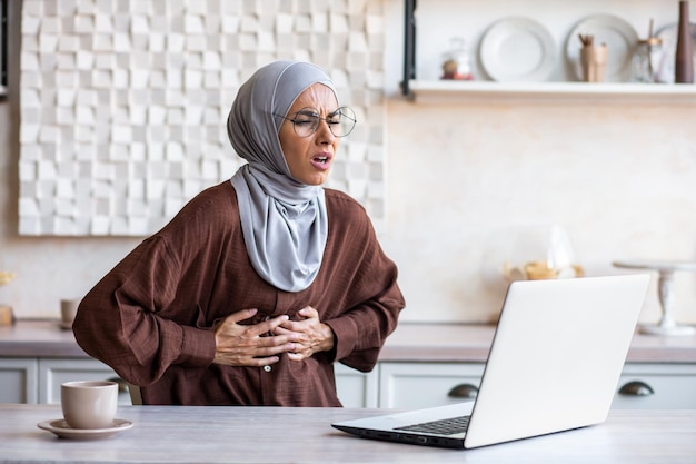 Bauchanfall einer jungen muslimischen Frau im Hijab, die zu Hause arbeitet und mit einem Laptop lernt