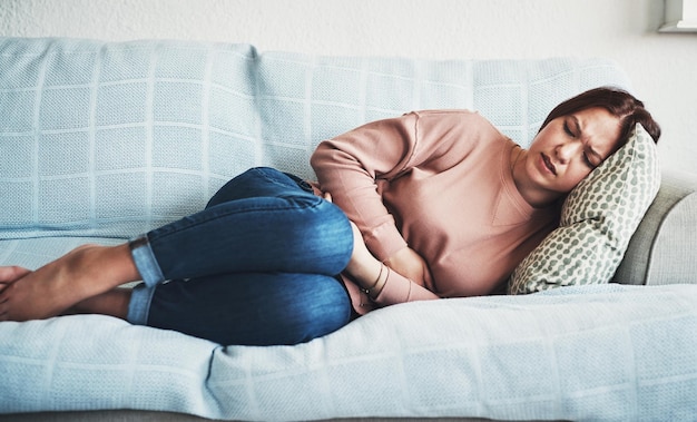 Bauchschmerzen oder Schmerzen einer Frau auf dem heimischen Sofa mit Menstruations- oder Regelkrämpfen im Wohnzimmer. Krankes Bauch- oder Dickdarmproblem einer weiblichen Person mit den Händen auf dem Bauch wegen Verstopfung, Verdauung oder Virus