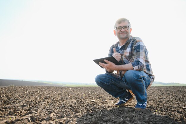 Bauer arbeitet im Frühjahr mit Tablet auf dem Feld. Ein älterer Bauer blickt auf Tablet und grüne Triebe. Ein kluger Agronom mit Tablet in der Hand überprüft das Feld. Umweltfreundliche Landwirtschaft