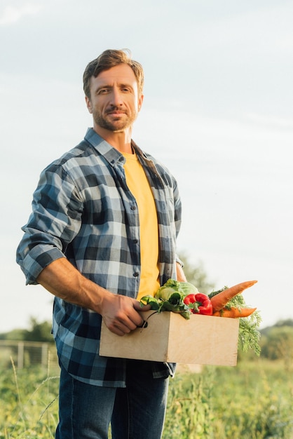 Bauer im karierten Hemd, der in die Kamera blickt, während er eine Kiste mit reifem Gemüse hält