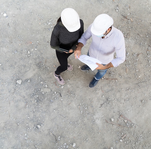 Bauherr und Ingenieur in der schützenden Gesichtsmaske arbeiten auf der Baustelle mit Blick auf Pläne Draufsicht