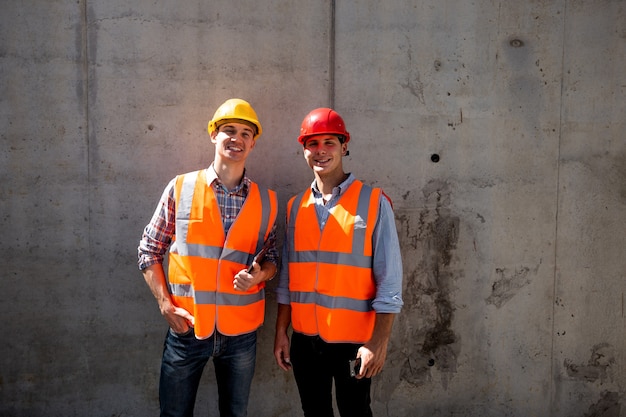 Bauingenieur und Architekt in orangefarbenen Arbeitswesten und Helmen stehen auf einem Betonwandhintergrund. .