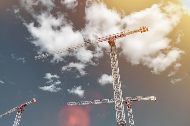 Baukräne arbeiten auf der Baustelle vor blauem Himmelshintergrund. Ansicht von unten auf den Industriekran. Konzept des Baus von Mehrfamilienhäusern und Renovierung von Wohnungen. Platz kopieren