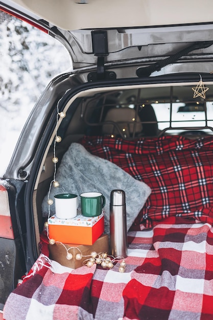 El baúl del auto está decorado para la celebración de Navidad y Año Nuevo en un bosque nevado de invierno Taza y termo para beber té caliente Acogedora tela escocesa roja y caja de regalo Picnic blanco en el bosque