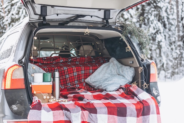 El baúl del auto está decorado para la celebración de Navidad y Año Nuevo en un bosque nevado de invierno Taza y termo para beber té caliente Acogedora tela escocesa roja y caja de regalo Picnic blanco en el bosque