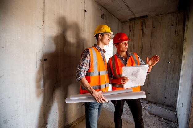 Bauleiter und Ingenieur in orangefarbenen Arbeitswesten und Helmen arbeiten mit der Baudokumentation innerhalb des im Bau befindlichen Gebäudes.