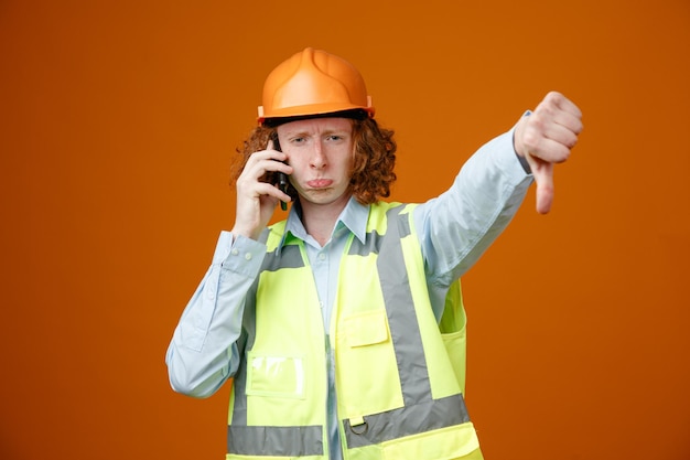 Baumeister junger Mann in Bauuniform und Schutzhelm telefoniert unzufrieden mit schiefem Mund und zeigt Daumen nach unten über orangefarbenem Hintergrund